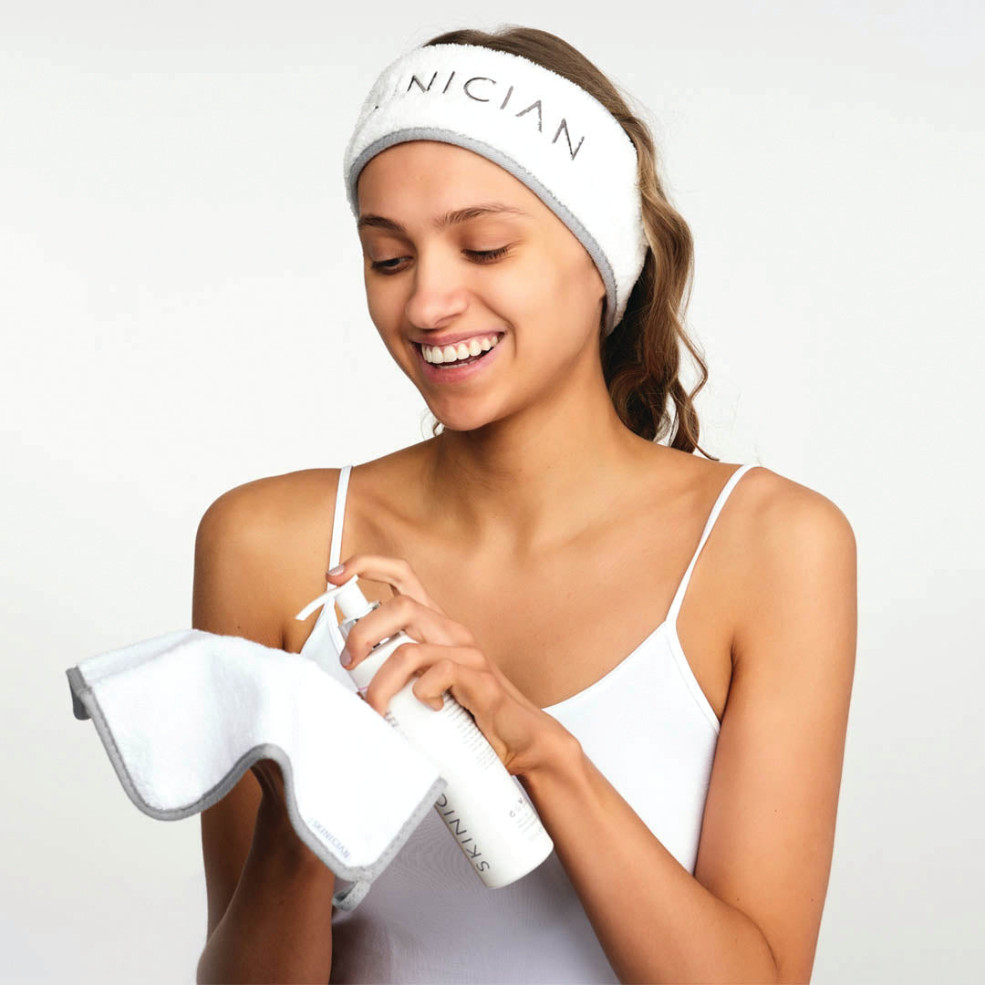 A smiling woman wearing a white headband with Skinician branding, holding a white face cloth
